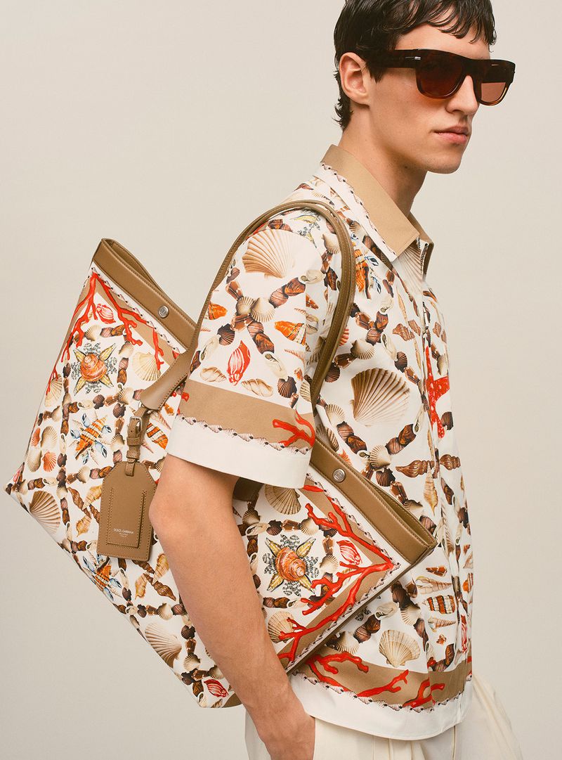 Man with light skin wearing a seashell-patterned shirt and sunglasses, holding a large tote bag with seashell and coral designs.