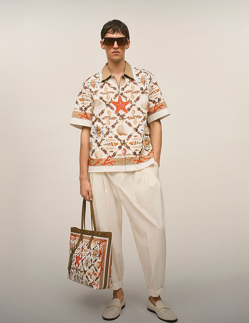 A fair-skinned adult male model is standing in a studio wearing a patterned shirt, pants, sunglasses and holding a tote bag.