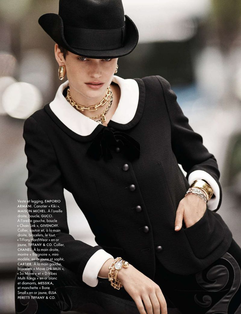 Portrait of a female fashion model wearing a black hat and black and white jacket, posing outdoors with gold jewelry and bracelets.