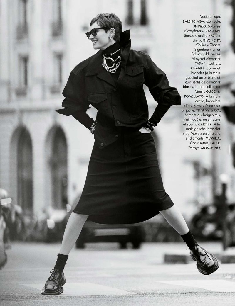 Black-and-white fashion shot of a woman in a black skirt suit and chunky shoes, smiling in front of a blurred urban background.