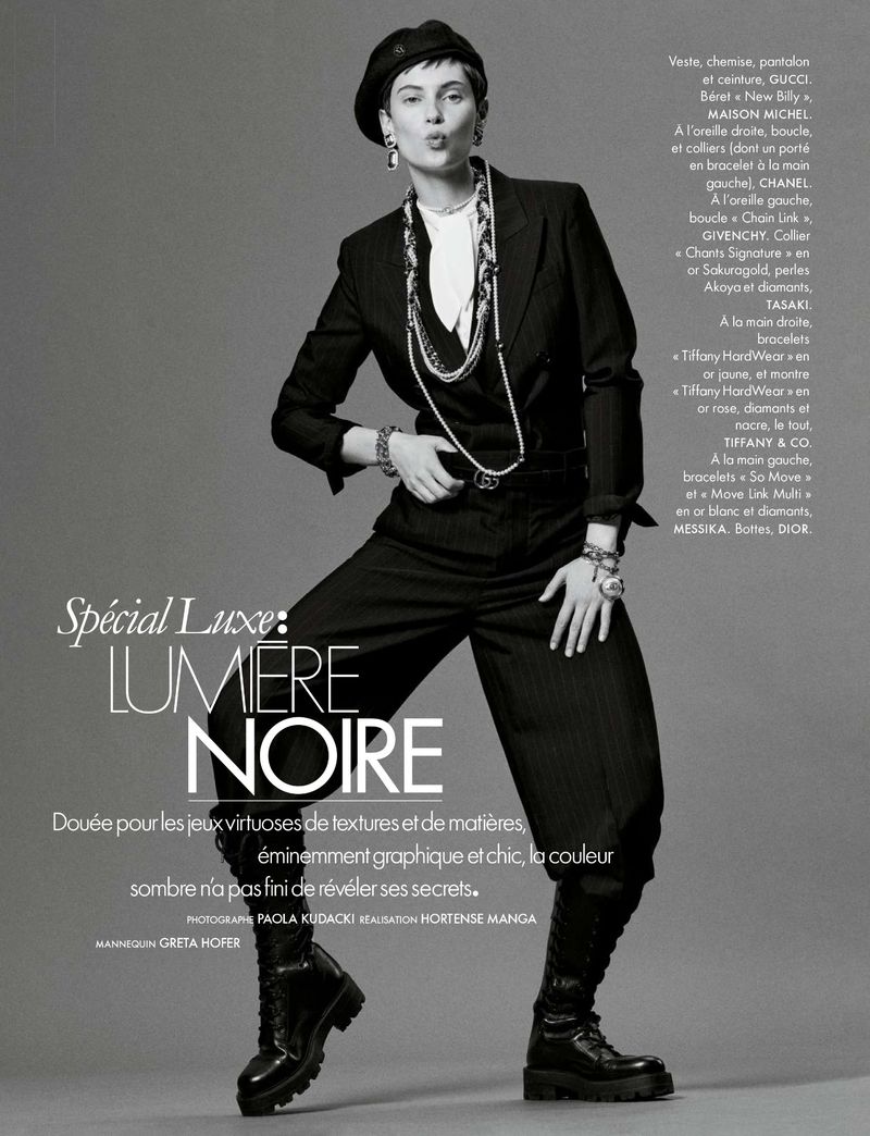 A woman in a black pantsuit and beret poses in a studio, adorned with layers of necklaces and bracelets, striking a chic pose.