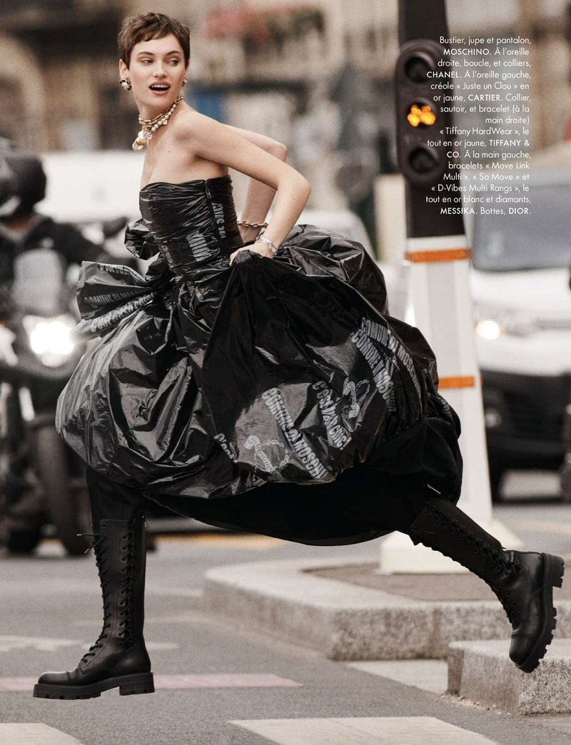 A model strides on a city crosswalk wearing a black strapless gown with knee-high boots, against a backdrop of blurred urban activity.