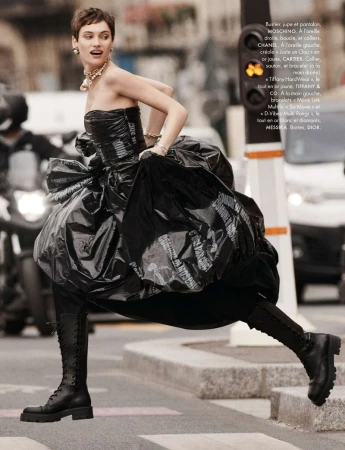 A model strides on a city crosswalk wearing a black strapless gown with knee-high boots, against a backdrop of blurred urban activity.