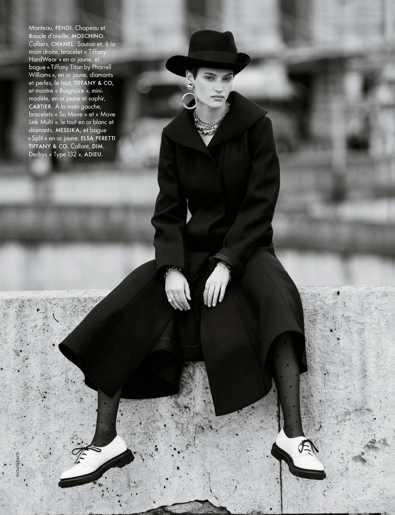 Monochromatic fashion shot features a model in a black coat, hat, and white shoes, seated on a concrete structure against an urban backdrop.