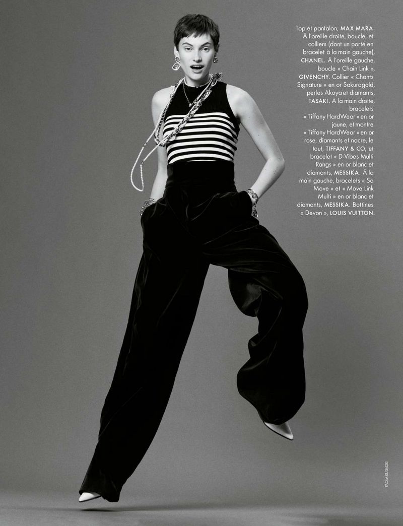 Fashion editorial shot of a woman in a striped top and black pants jumping, accessorized with statement jewelry, against a grey background.