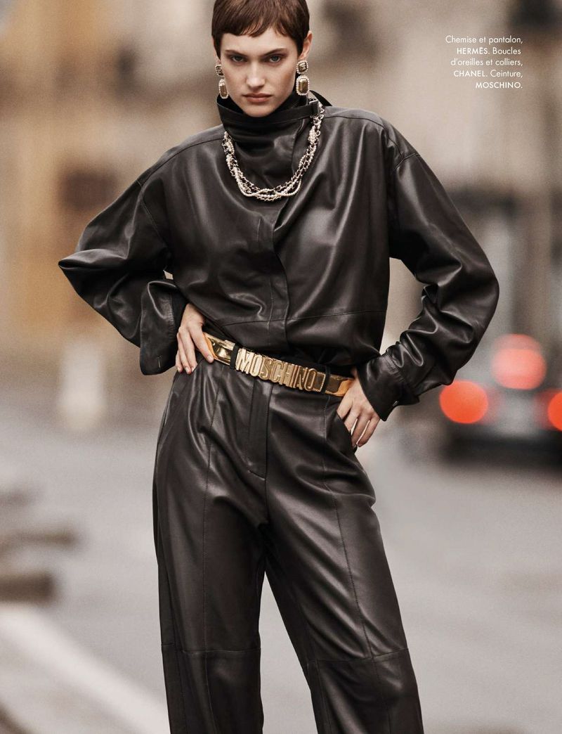 A woman in black leather top and pants with a gold belt poses on a city street with blurred cars in the background for a fashion editorial.