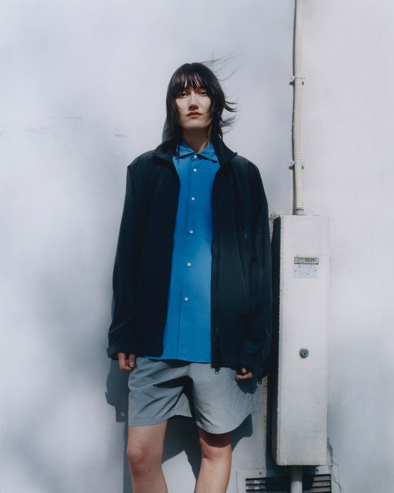 Fashionable person posing in a navy jacket, blue button-down shirt, and grey shorts standing against a white wall outdoors.