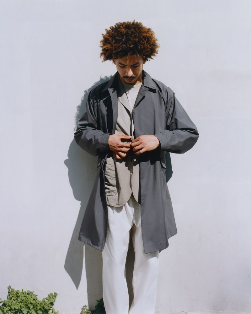 Man in a grey trench coat against a white wall, captured in a medium shot with natural lighting, showcases fashion and style.