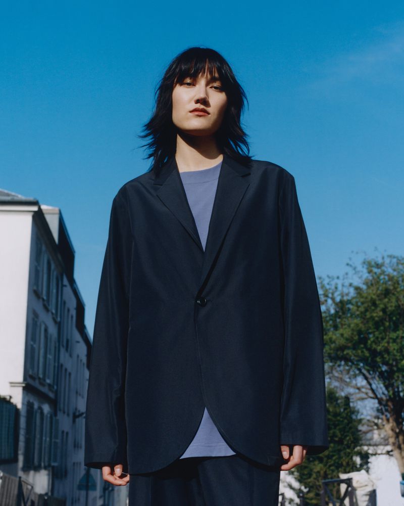 Outdoor portrait of adult, fair-skinned person with dark hair wearing navy blazer in an urban setting, under clear blue sky.