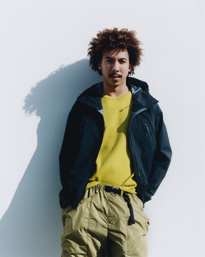Man in a blue hooded jacket and khaki pants stands against a white wall with his shadow behind him, creating a relaxed fashion portrait.