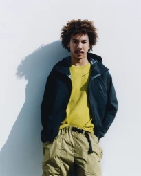 Man with curly hair wearing a dark jacket over a yellow shirt and khaki pants against a plain white wall.