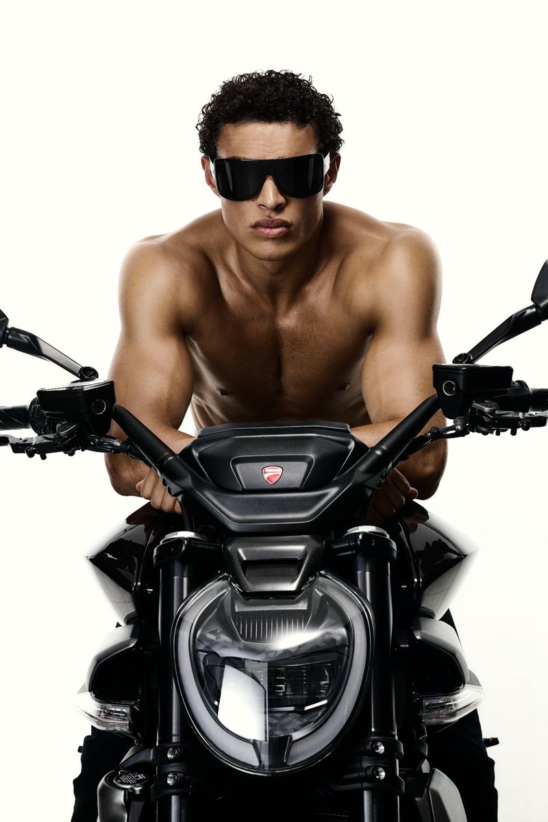 Shirtless man with sunglasses poses on a black Ducati motorcycle against a white background in a studio shot for automotive lifestyle.