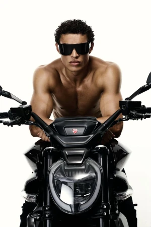 Shirtless man with sunglasses poses on a black Ducati motorcycle against a white background in a studio shot for automotive lifestyle.