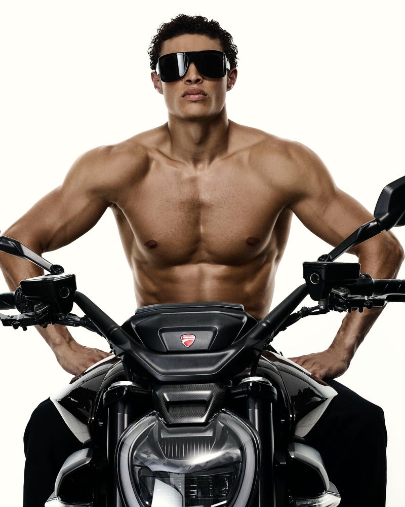 Shirtless muscular man poses confidently on a black and white Ducati motorcycle against a bright white background, wearing dark sunglasses.