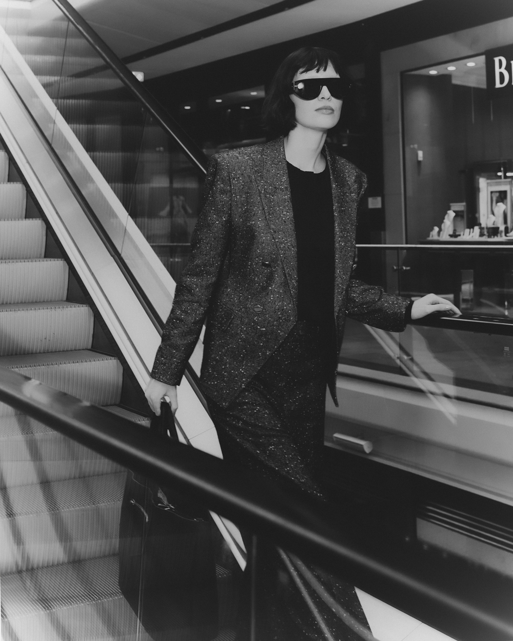 Black and white fashion shot: a woman in a tailored suit and sunglasses stands on an escalator holding a briefcase. Shop display visible.