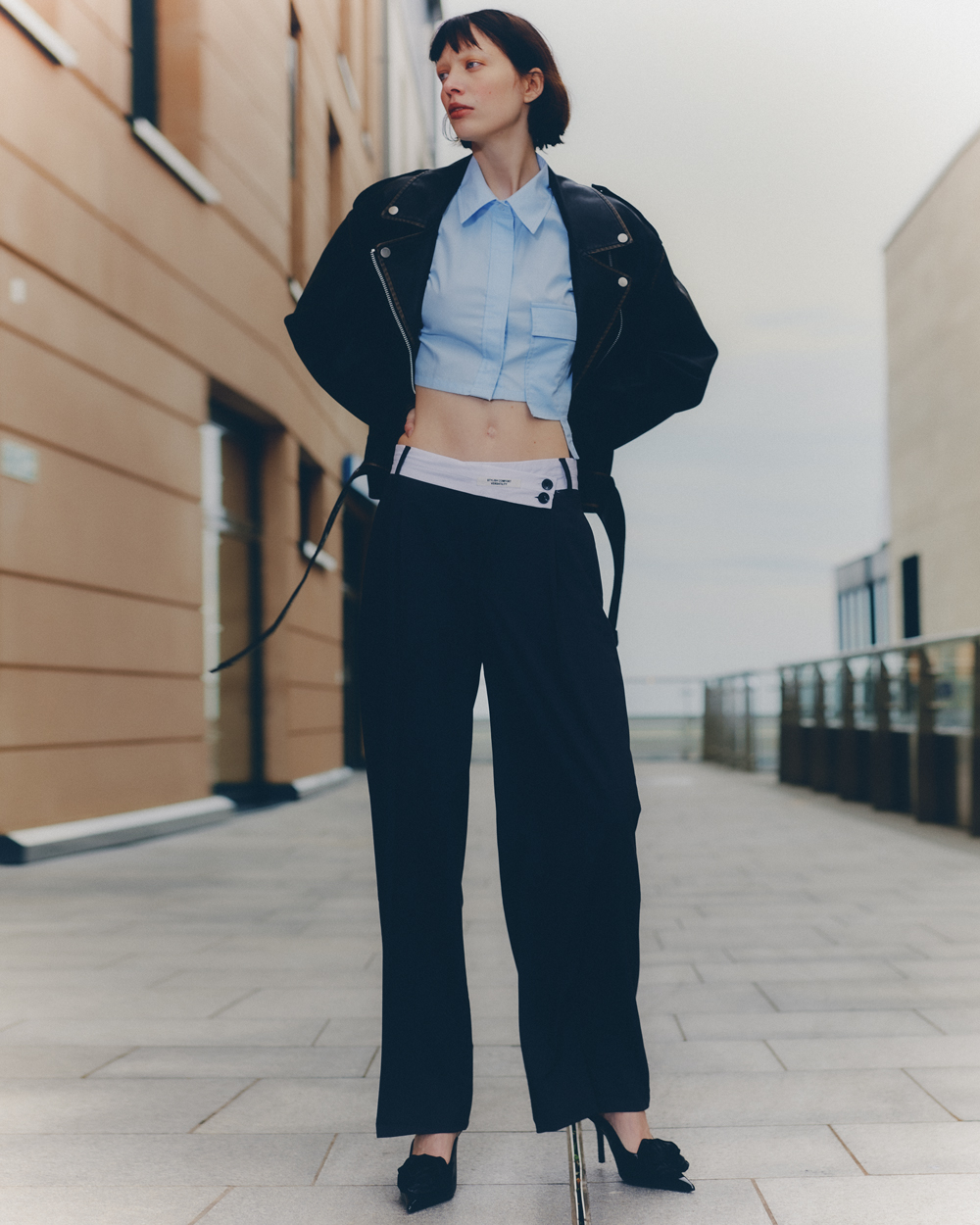 Full-length shot of a slender woman in a blue crop top, black jacket, and wide-leg pants, posing outdoors against a brick building.