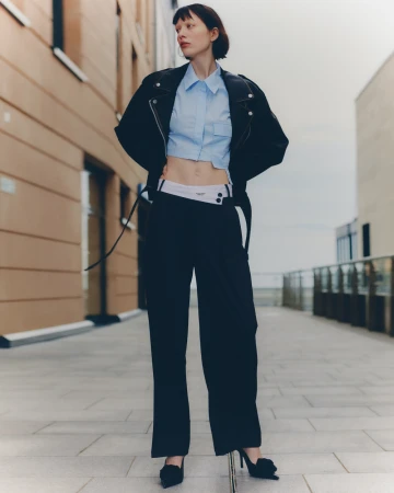 Full-length shot of a slender woman in a blue crop top, black jacket, and wide-leg pants, posing outdoors against a brick building.