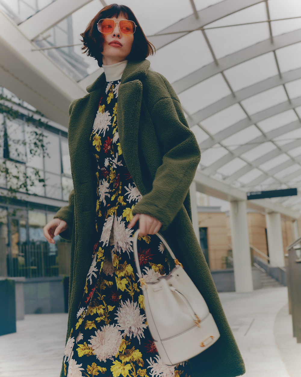 Fashionable adult woman in floral dress and olive coat walks in an urban setting carrying a white handbag, with a modern building backdrop.