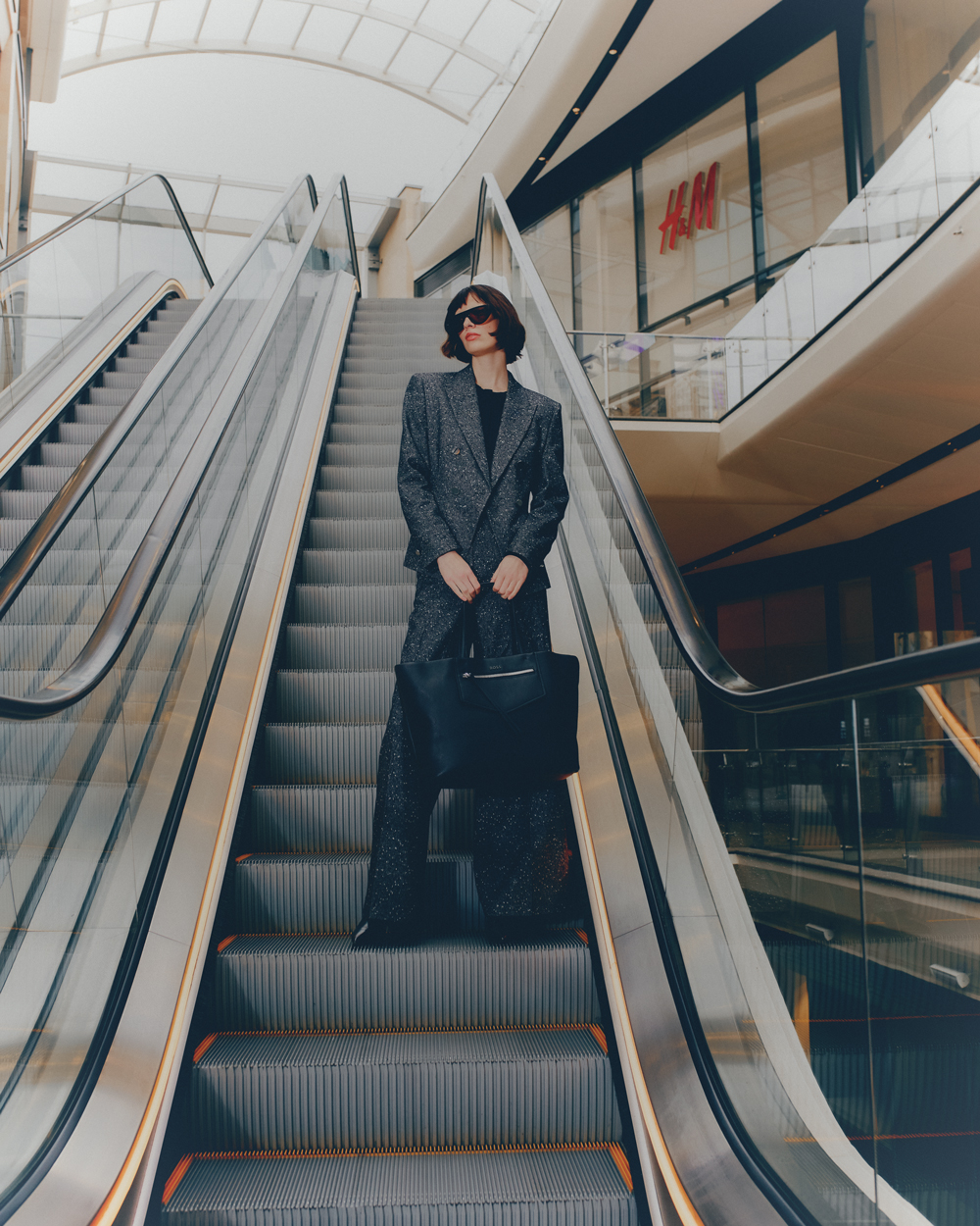 Woman in patterned suit on escalator in shopping mall, holding black purse, wearing sunglasses, H&M store background, indoor setting.