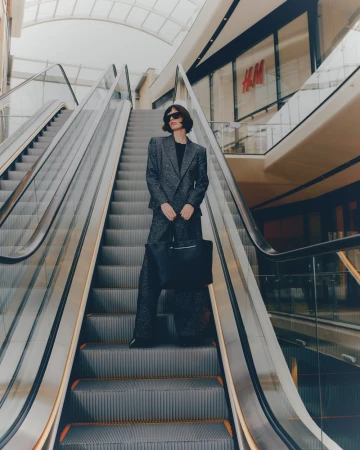 Woman in patterned suit on escalator in shopping mall, holding black purse, wearing sunglasses, H&M store background, indoor setting.