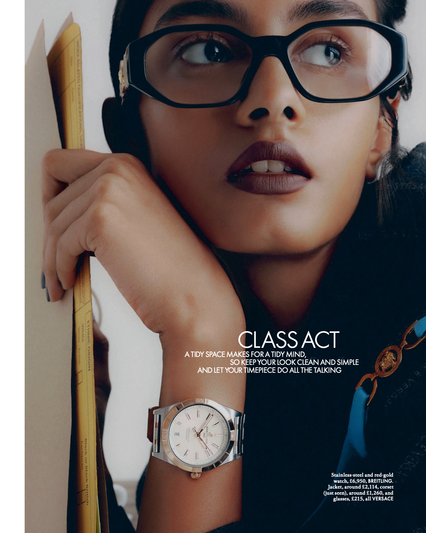 Close-up studio shot of a woman with dark hair and black glasses, wearing a watch, holding a folder, and looking off into the distance.
