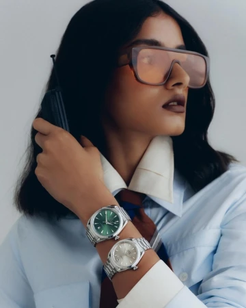 Woman with oversized glasses and a shirt wearing two silver wristwatches. Product photography of a watch with a green face and a silver face.