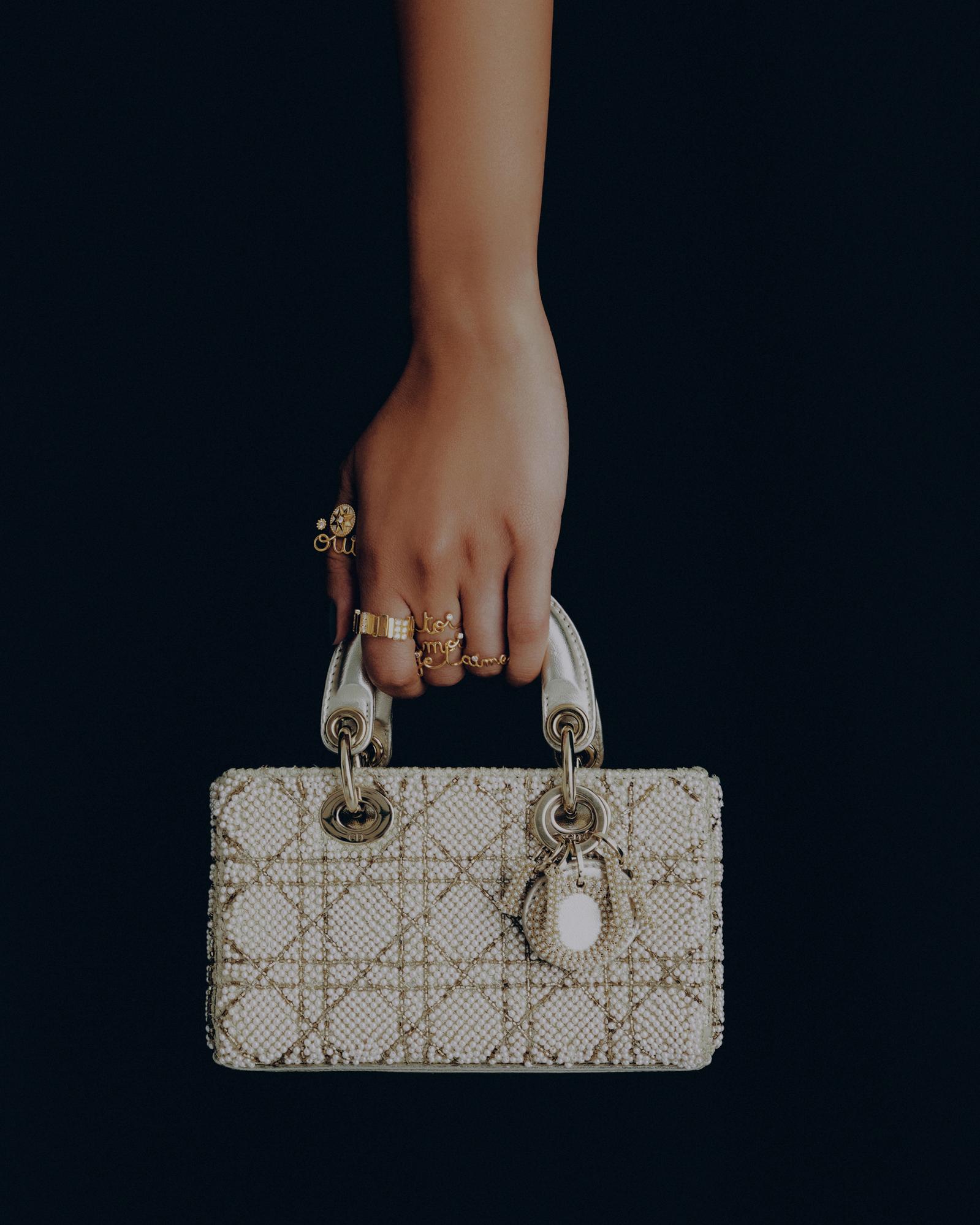 A hand holds a white, beaded Lady Dior bag against a black background. The hand has several gold rings. Fashion product shot.