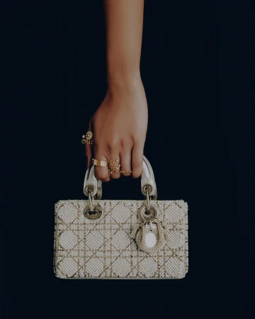 A hand holds a white, beaded Lady Dior bag against a black background. The hand has several gold rings. Fashion product shot.