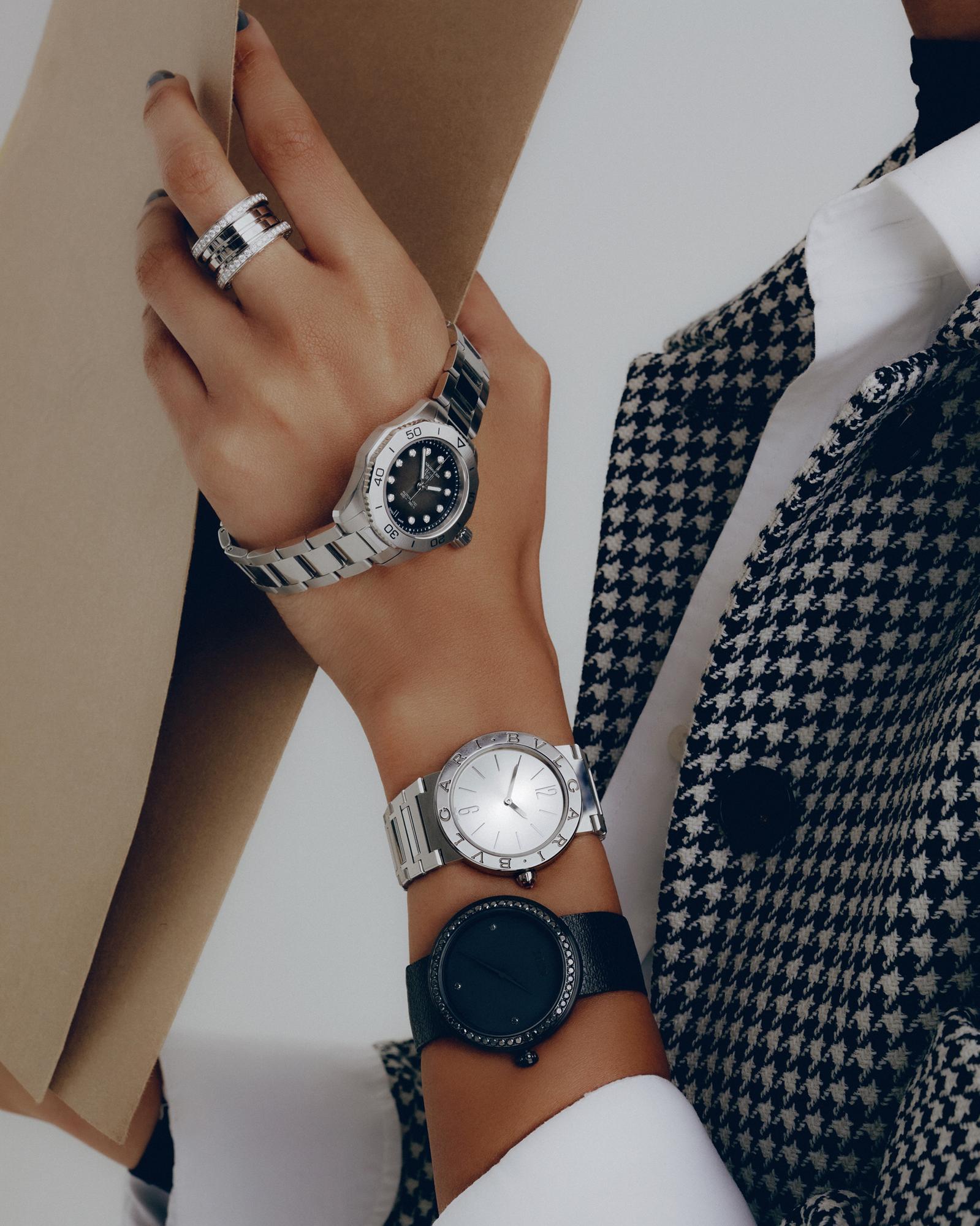 Close-up of a person's wrists wearing multiple wristwatches and a ring against a houndstooth patterned jacket and tan fabric.