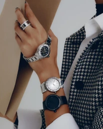 Close-up of a person wearing three luxury wristwatches and a diamond ring while holding a beige folder, dressed in a houndstooth blazer.