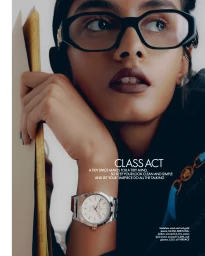 Close-up portrait of a woman wearing black geometric glasses, holding yellow documents, with a luxury watch on her wrist.