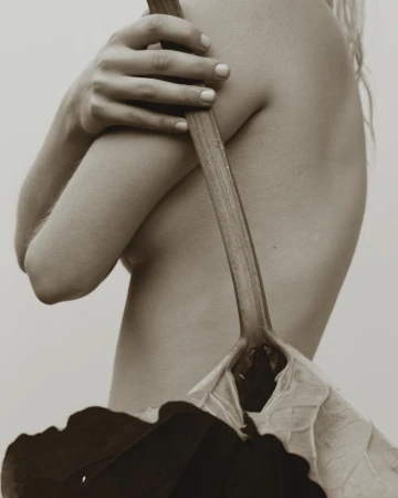 Monochrome image of a partially nude woman holding a large green leaf and stem, posing against a light background.