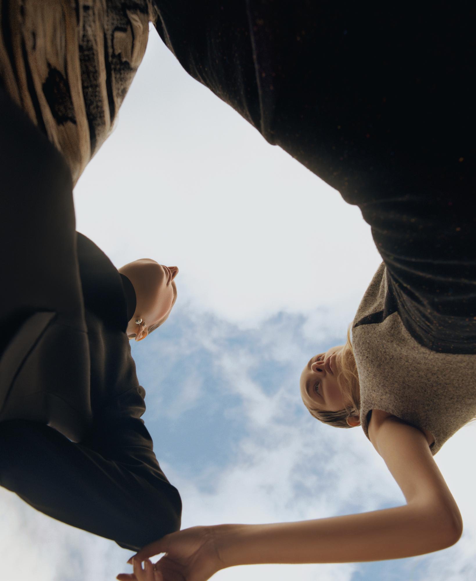Low angle shot of two people standing close together under a cloudy sky, wearing dark casual clothing, their faces tilted towards the camera.