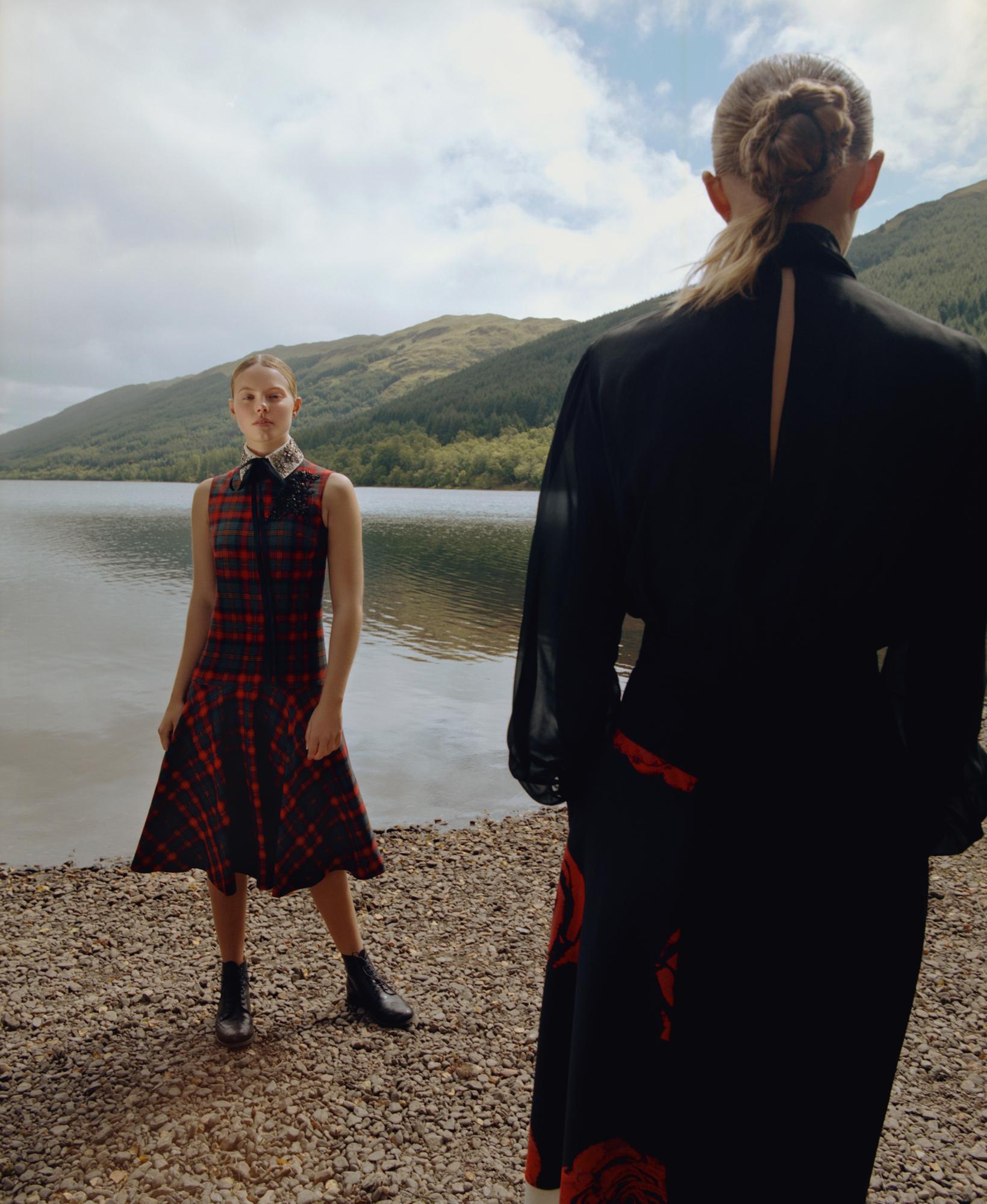 Two fair-skinned women pose at a lakeside, one wearing a plaid dress and ankle boots, the other wearing a long black dress with red floral detail.