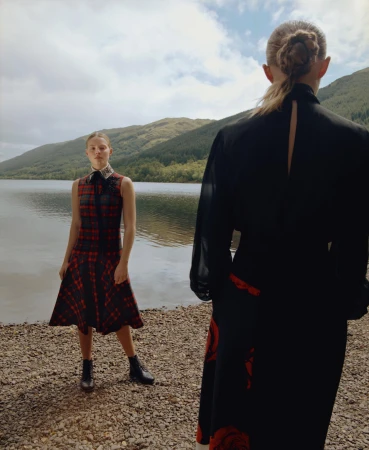 Two fair-skinned women pose at a lakeside, one wearing a plaid dress and ankle boots, the other wearing a long black dress with red floral detail.