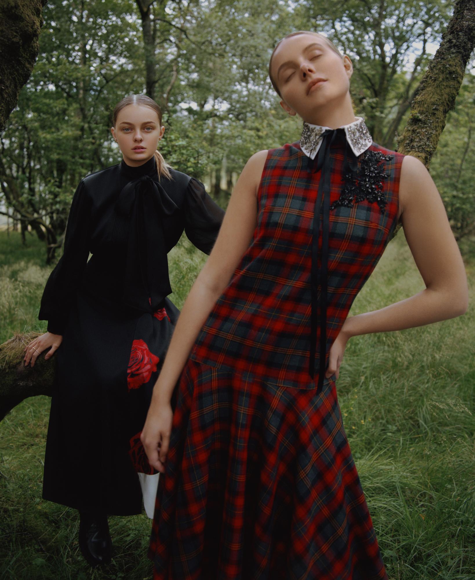 Two adult female fashion models pose outdoors wearing dresses with plaid and floral patterns in a wooded environment during daytime.
