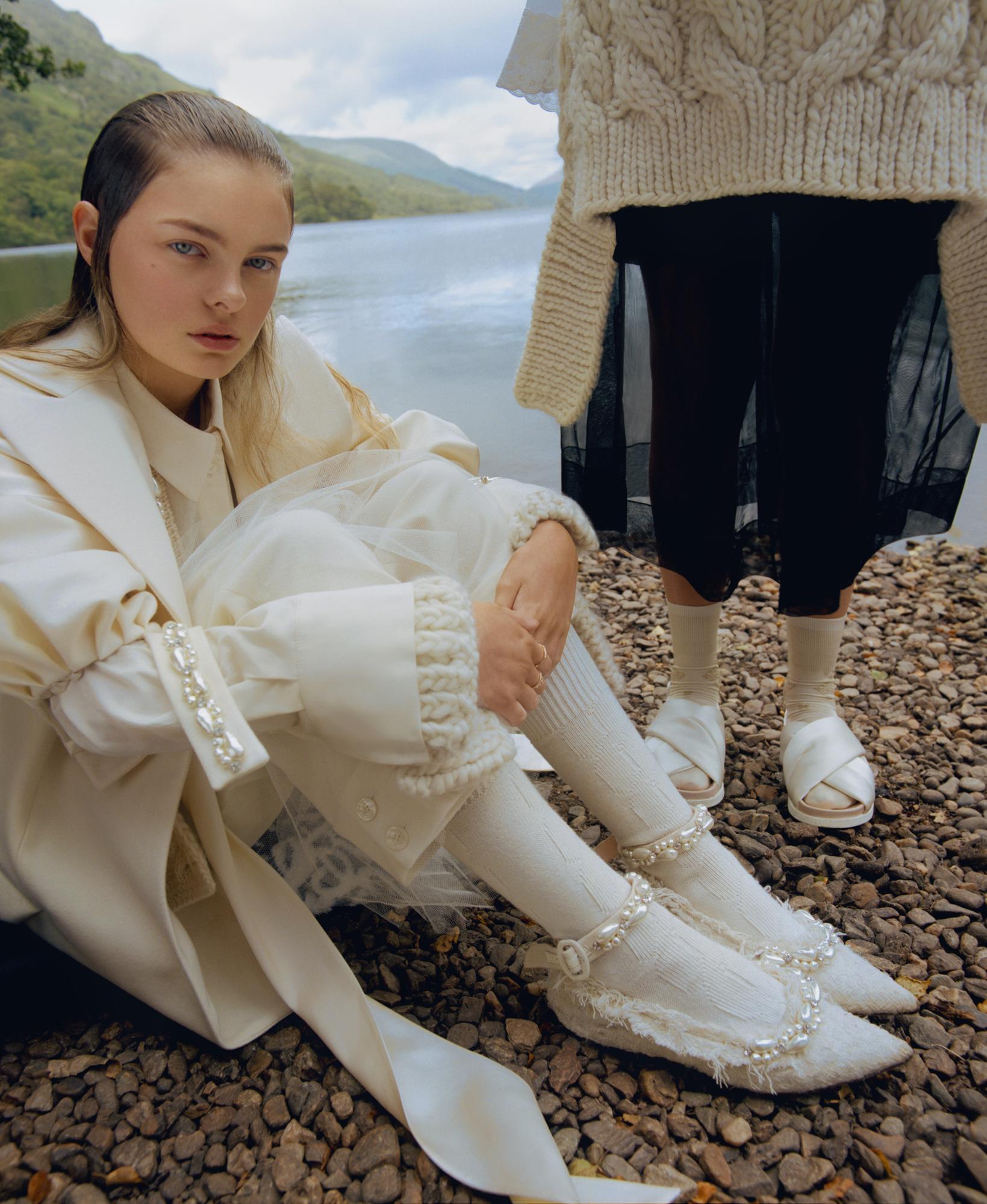 Two women pose in matching off-white outfits by a lake with mountains in the distance, showcasing fashion and nature.