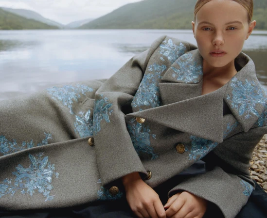 Woman in grey floral coat poses by lake with mountains in background. Fashion shot with natural light and shallow depth of field.