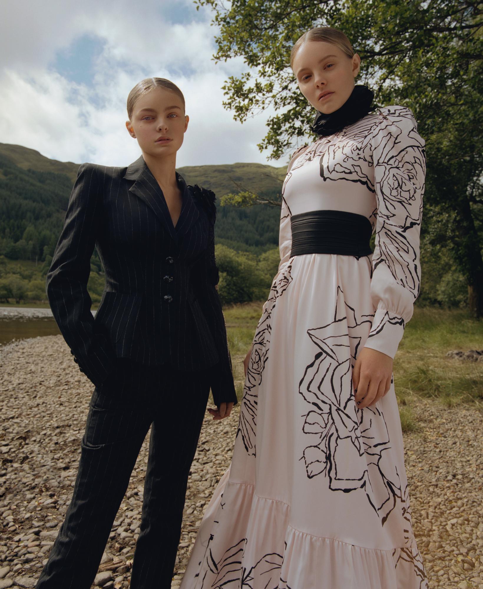Two fashion models in designer clothing pose outdoors by a river with a mountain backdrop, under a cloudy sky for a fashion editorial.