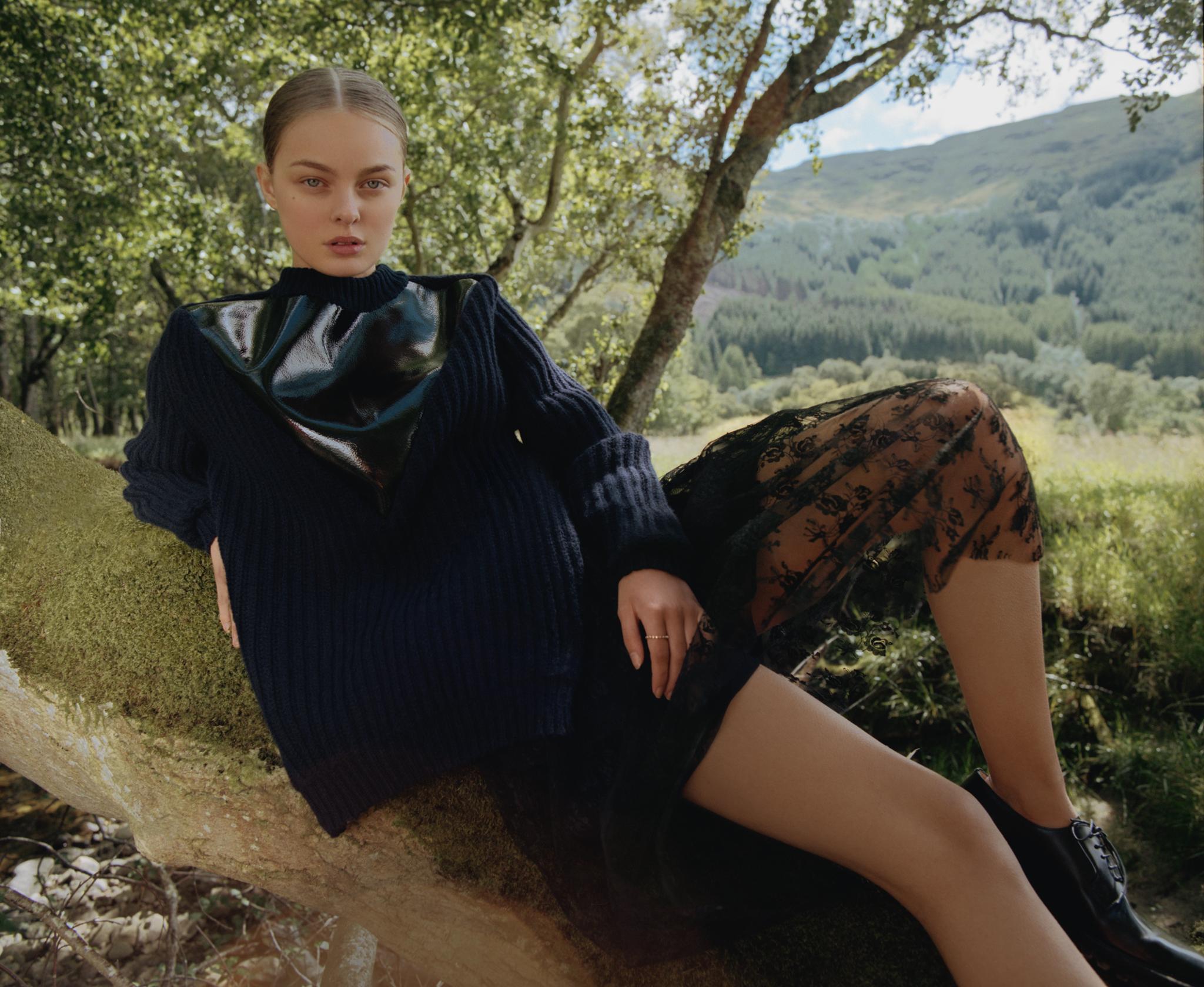 Adult female model poses on tree in fashion editorial, wearing black top and lace skirt, rural outdoor landscape, medium shot.