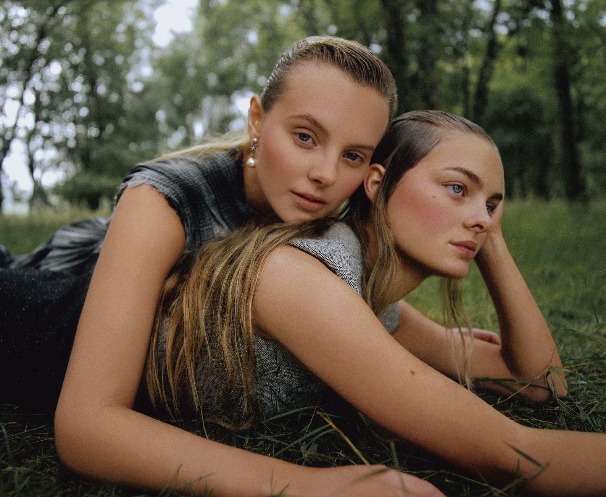 Two young women with light complexions are laying in the grass in a natural outdoor environment surrounded by green foliage and trees.
