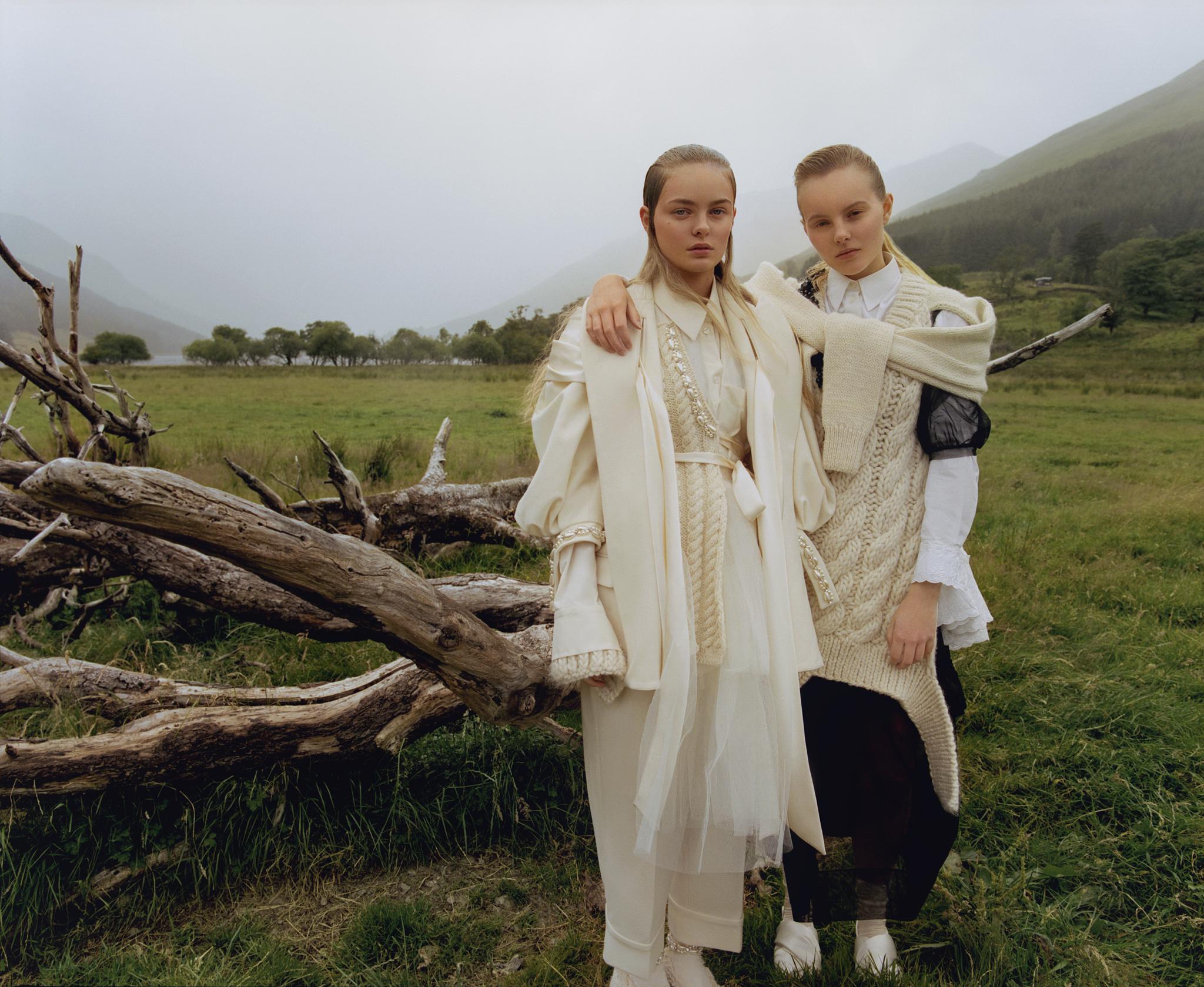 Two fair-skinned adult women pose outdoors on a grassy field wearing cream-colored fashion against a background of mountains and fog.