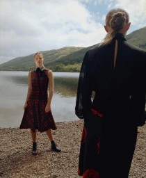 Two women by a lakeside on a pebbled shore with green hills under an overcast sky, one in a plaid dress and one in a black top.