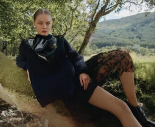 Woman in dark knit sweater and lace skirt reclining on a mossy tree trunk in an outdoor woodland setting.