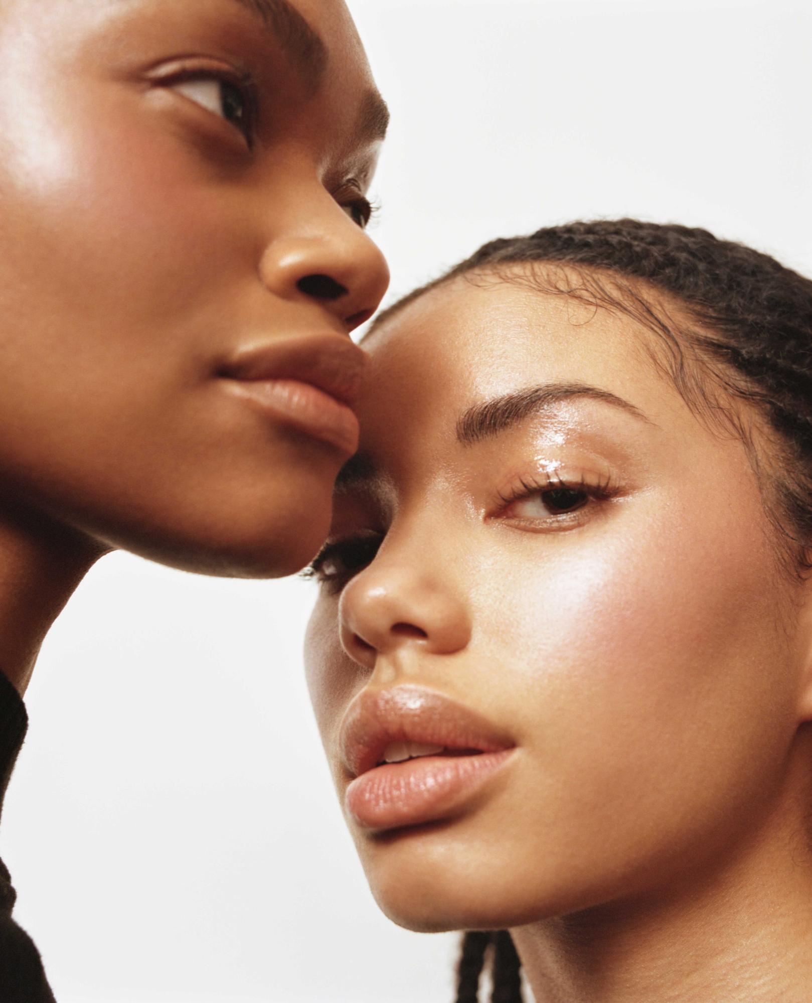 Close-up portrait of two dark-skinned women with glossy skin, posing together in a studio with a seamless white background. Beauty shot.