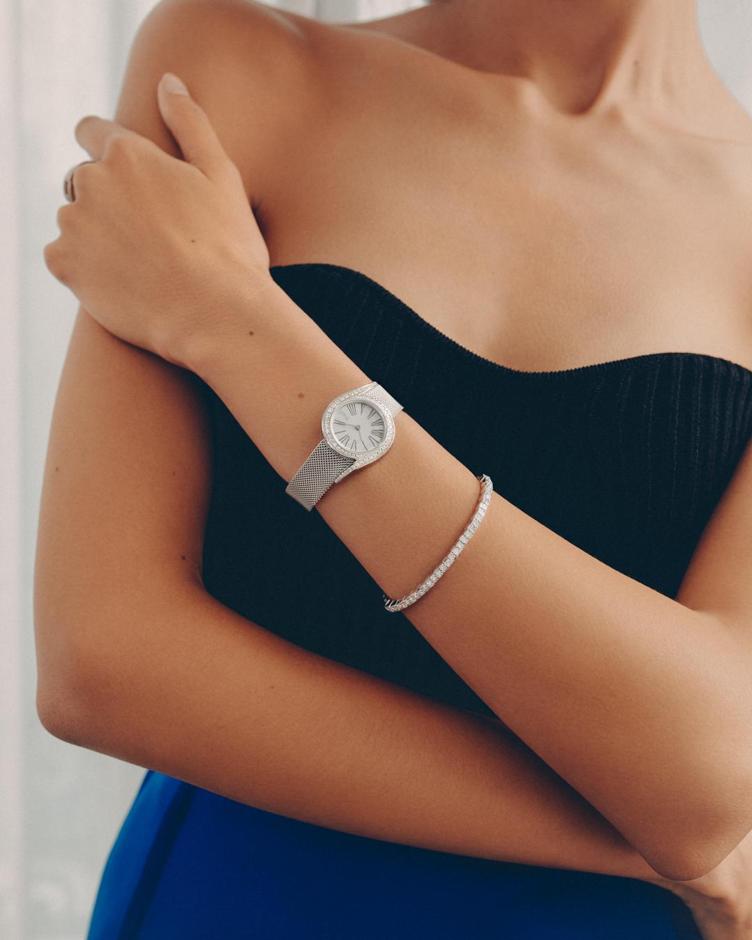 Elegant indoor shot of a woman showcasing a silver wristwatch and diamond bracelet, wearing a black strapless top and blue bottom.