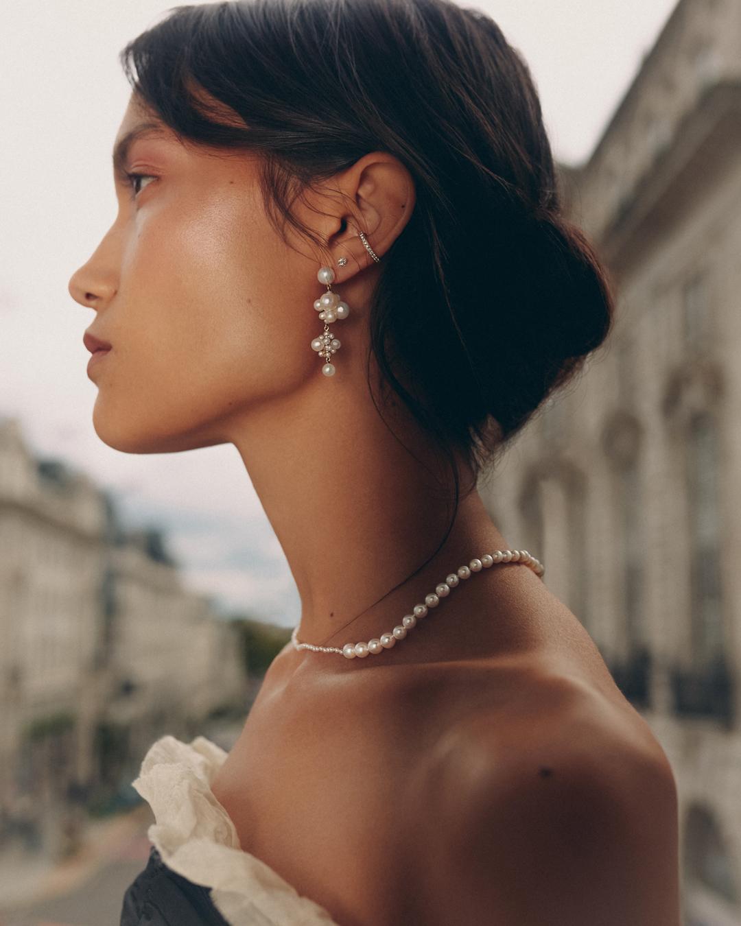 Profile of a woman wearing a pearl necklace and earrings, set against a backdrop of historic buildings beneath a cloudy sky.