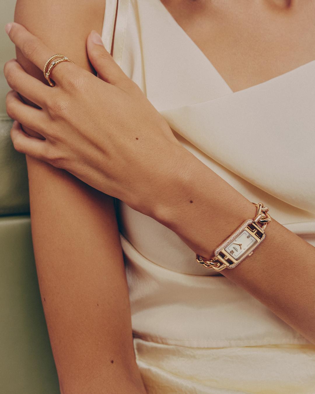 Close-up of a woman's wrist with a gold chain watch, an elegant gold ring, and a white dress. Soft lighting in an indoor setting.