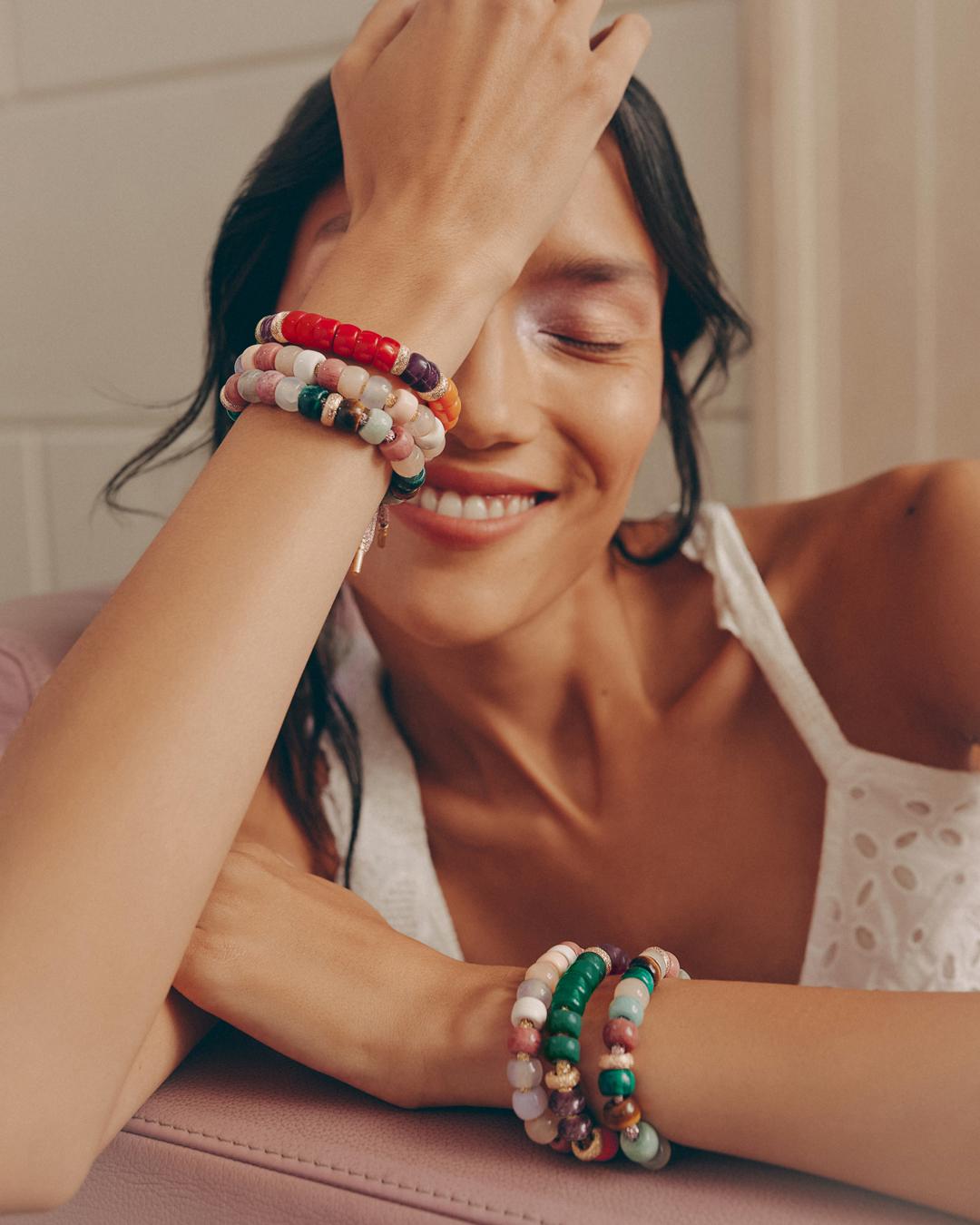 Smiling woman with dark hair wearing beaded bracelets on both wrists, leaning on a pink surface, indoors, with natural light.