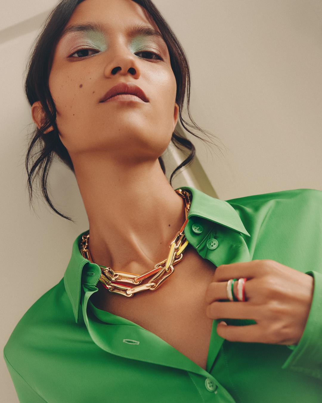 Woman with light green eye makeup poses wearing a green button-down shirt, a chunky gold necklace, and multi-colored rings on her finger.